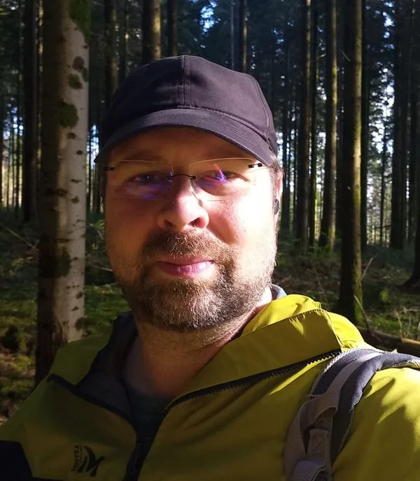 Kévin Solinski en forêt, casquette et veste de randonnée, souriant.